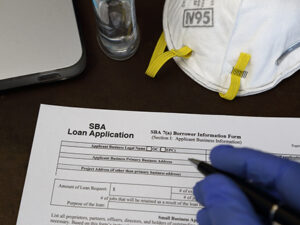 A rubber gloved hand is shown about to fill out a Small Business Administration aka SBA loan application form, with an N95 respirator dust mask and antibacterial gel nearby.