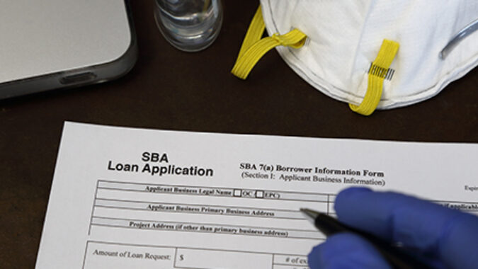 A rubber gloved hand is shown about to fill out a Small Business Administration aka SBA loan application form, with an N95 respirator dust mask and antibacterial gel nearby.