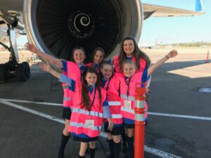 Five school girls and one adult wearing pink safety vests standing in front of a commercial airplane's jet engine on the tarmac